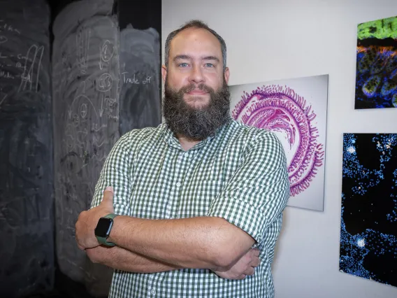 Portrait of a man in a checked shirt standing with his arms crossed in front of artwork. 