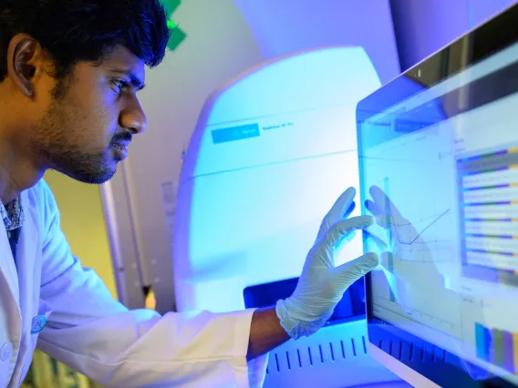 A graduate student in a white lab coat touches a computer monitor while looking at it intently. 