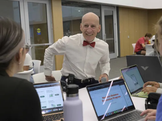 Dr. Paul Gordon smiles as he leans on a table in a classroom and talks with students who are on laptops. 