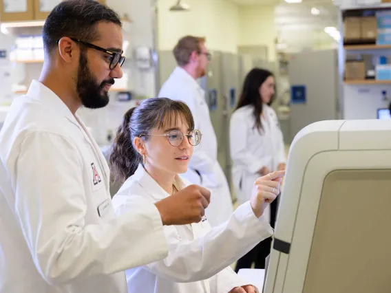 Two graduate students in white lab coats work on a large machine in a laboratory. 