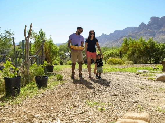 A family of three walks outdoors in a green desert environment with mountains in the background.