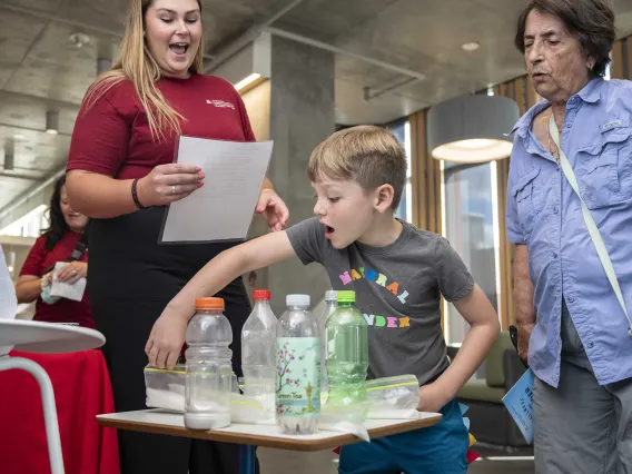 A surprised young boy lifts a bag of sugar behind soda bottles on a table while two women watch him. 