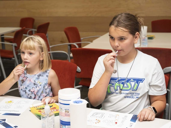 Two young girls sit at a table while they taste small pieces of flavored paper. 