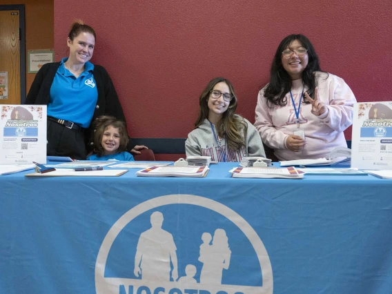 Three adults and a child stand behind a blue information table with some handouts and small posters. 