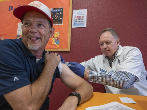 A man sitting at a table looks away and sticks out his tongue as a medical working gives him a shot in the arm.