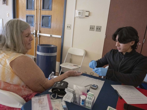 A woman sits at a table with her hand extended for a blood test being administered by a person wearing medical gloves. 