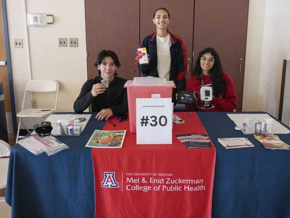 Three university students stand ready to give information at a table with a sign that reads “Mel and Enid Zuckerman College of Public Health.”