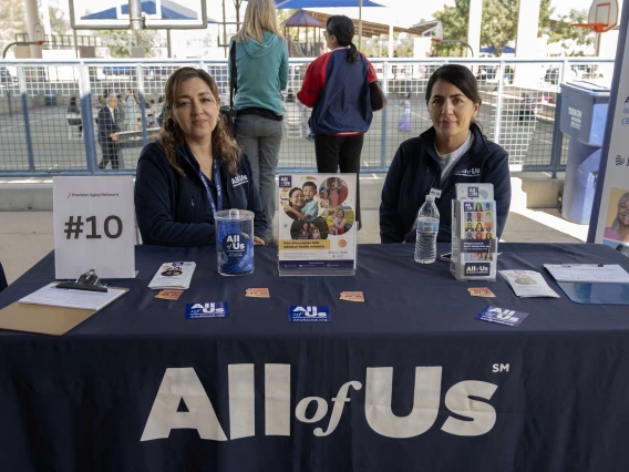 Two women stand behind an information table with a large sign that reads “All of Us.”