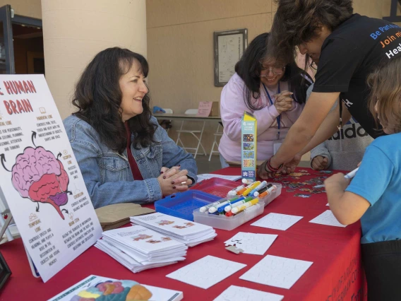 A woman sits at an information table covered with brochures and posters as people stand and talk with her. 