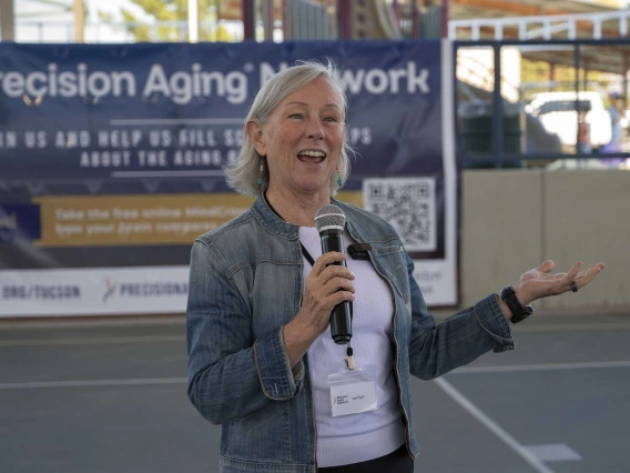 A woman stands on an outdoor sports court holding a microphone and speaking. She is smiling and holding up one hand as she speaks. 