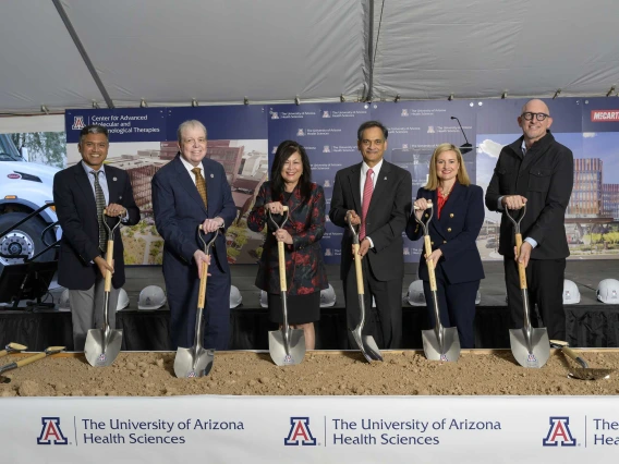 Six smiling people in suits and dresses hold shovels in sand with a University of Arizona Health Sciences banner in front of them.