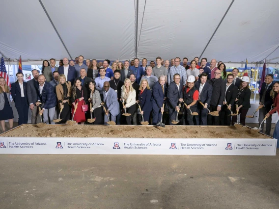 Several dozen people stand behind a large box of sand with shovels in hand. The banner across the front of the box reads “University of Arizona Health Sciences.”