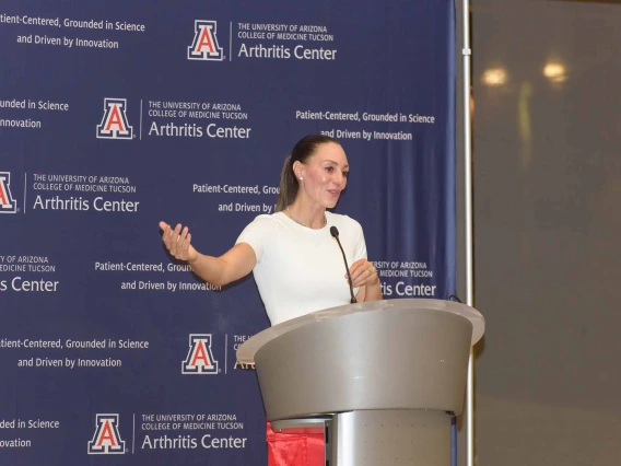 University of Arizona women’s basketball coach Adia Barnes speaks at a lectern in front of a blue backdrop with U of A Arthritis Center logos on it.