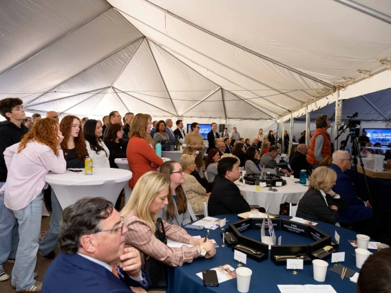 Hundreds of people sit and stand under a large white tent while listening to a speaker. 