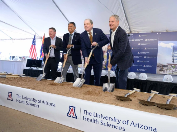 Four smiling men in suits hold shovels in sand with a University of Arizona Health Sciences banner in front of them.