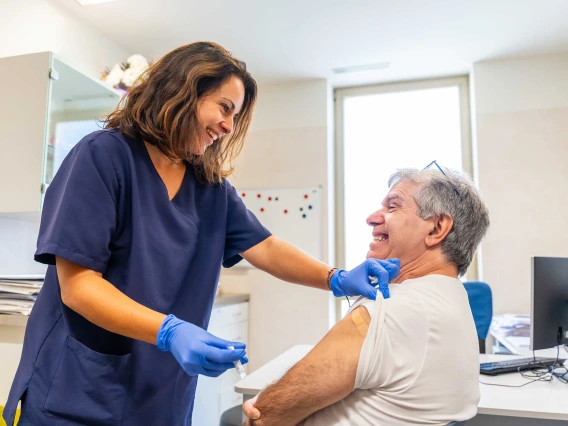 A female health worker chats with a seated male patient after administering a vaccine. Both are smiling.