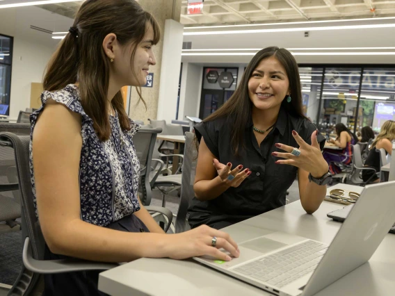 Photo of a young woman with her hand on a lap top who is looking at Caleigh Curley as Curley gestures with her hands. 