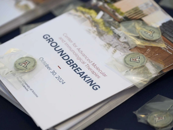 A stack of event programs with small, silver lapel pins scattered around it sits on a table. The program cover reads “Groundbreaking.” 