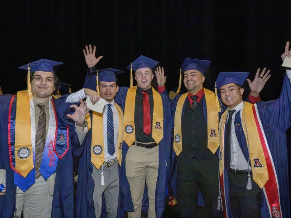 Five male nursing students wearing caps and gowns smile and wave as they stand side by side.