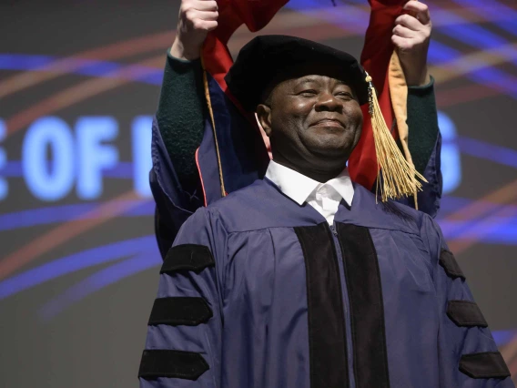 A Doctor of Nursing Practice student wearing a graduation cap and gown smiles as an unseen professor places a hood over his shoulders. 
