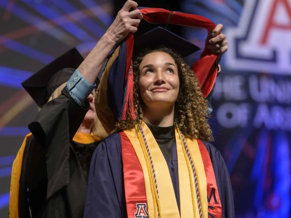 A woman wearing graduation regalia looks up and smiles as a traditional graduation hood is placed over her shoulders by a professor standing behind her. 