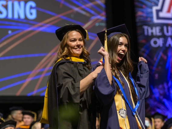 A young women in a graduation cap and gown pumps her arms in the air and smiles as a professor puts a ribbon around her neck