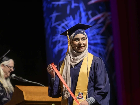 A woman wearing a hijab and traditional graduation regalia smiles as she walks across a stage. 