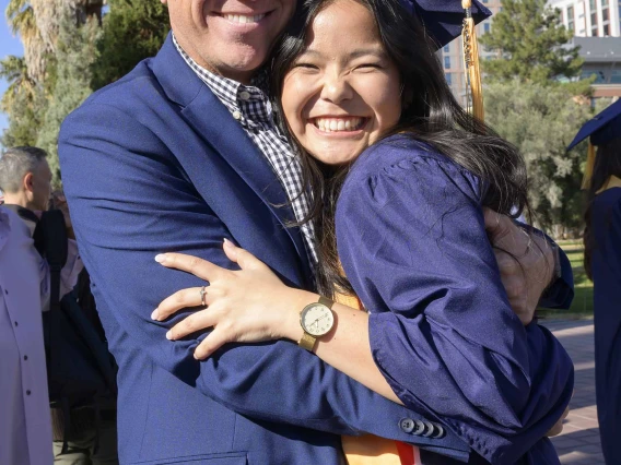 A young women dressed in a graduation cap and gown smiles as she hugs her smiling father.
