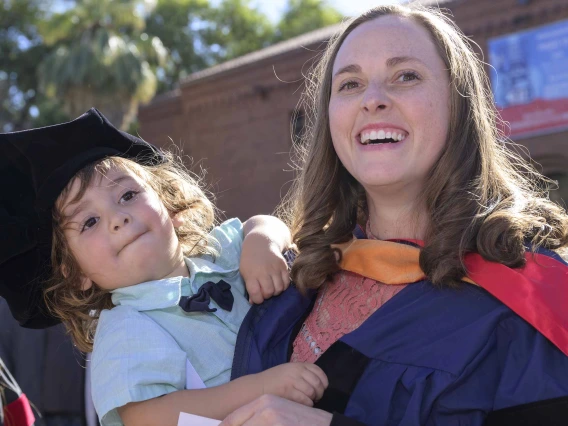 A smiling woman wearing a graduation gown holds a child who is wearing her graduation cap outside. 