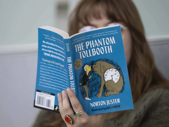 Close-up photo of the book “The Phantom Tollbooth” being help up and read by a person who is blurred into the background