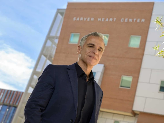 A man in a dark suit coat and black shirt stands outside a building that has Sarver Heart Center signage.