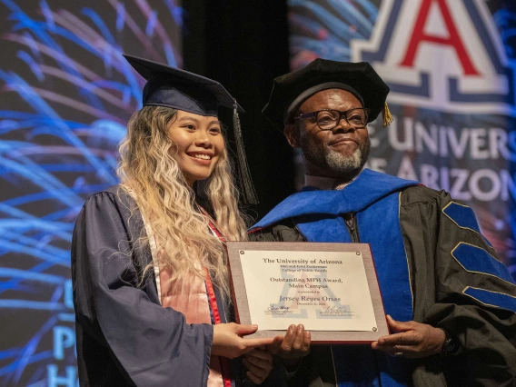 A student and a professor wearing graduation regalia stand next to each other, smiling and holding a certificate. 