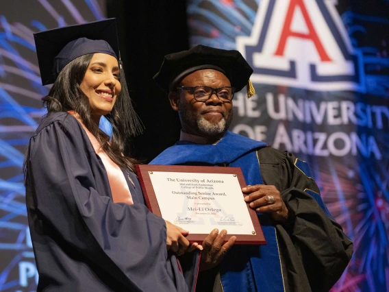 A student and a professor wearing graduation regalia stand next to each other, smiling and holding a certificate.