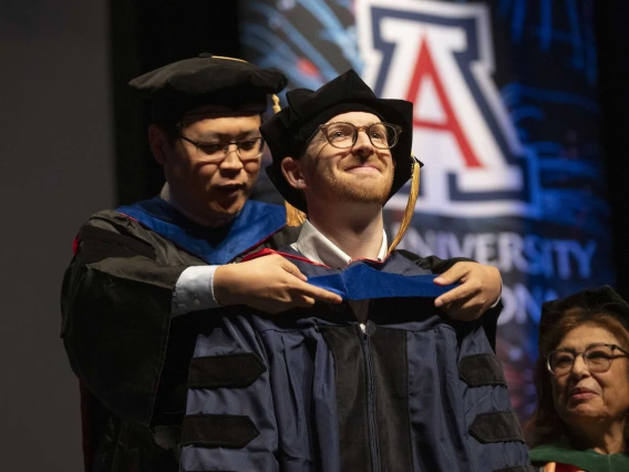 A professor places a sash on the shoulders of a graduate. Both are wearing caps and gowns. 