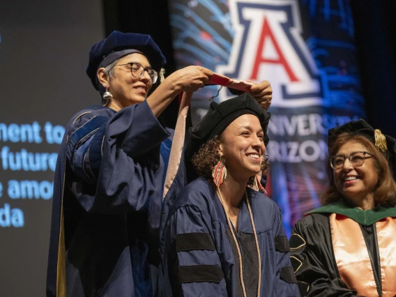 A professor places a sash on the shoulders of a graduate. Both are wearing caps and gowns.