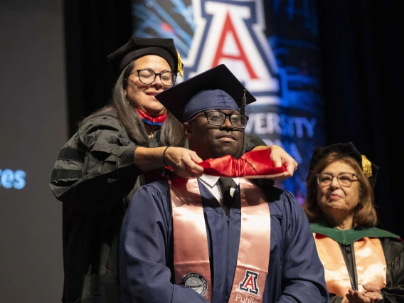 A professor places a sash on the shoulders of a graduate. Both are wearing caps and gowns.