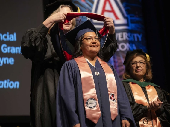 A professor places a sash on the shoulders of a graduate. Both are wearing caps and gowns.