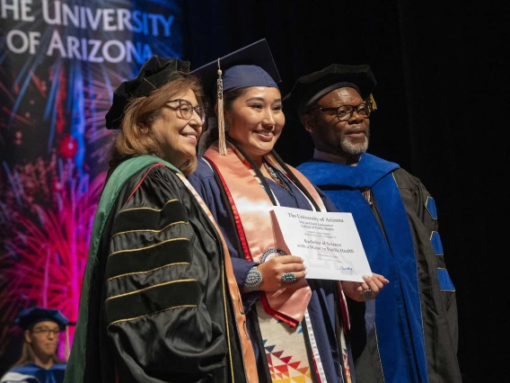 Professors stand on each side of a graduate, who is holding a certificate. All are wearing graduation caps and gowns. 