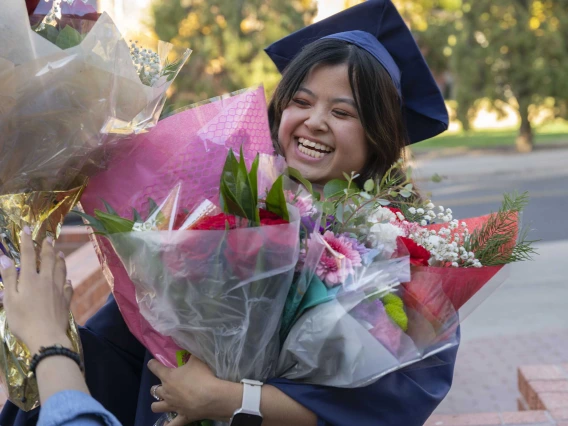 A smiling graduate stands outside with her arms full of flowers. 