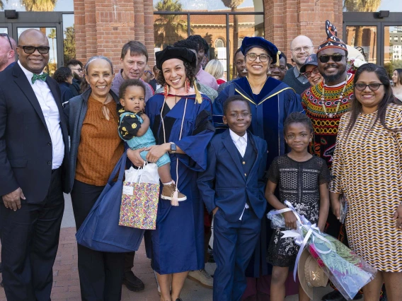 A graduate wearing a cap and gown holds a child while standing with family and friends outside after her graduation ceremony.