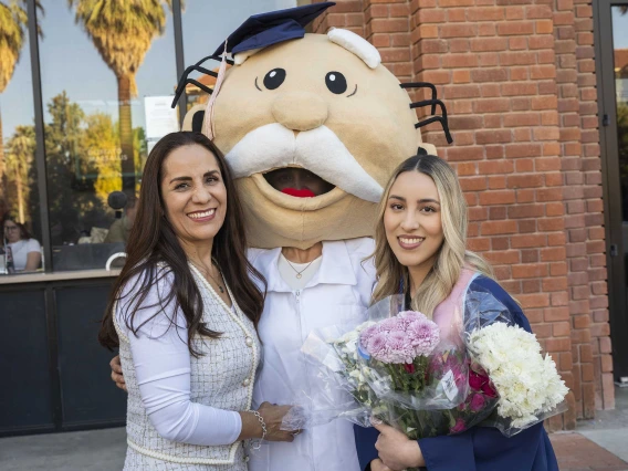 A graduate wearing a cap and gown smiles as she holds flowers while standing next to her mom and her sister, who is wearing a large mascot head adorned with a graduation cap. 