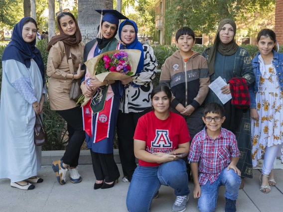 A graduate wearing a cap and gown and holding flowers stands with eight members of her family outside after her graduation ceremony. 