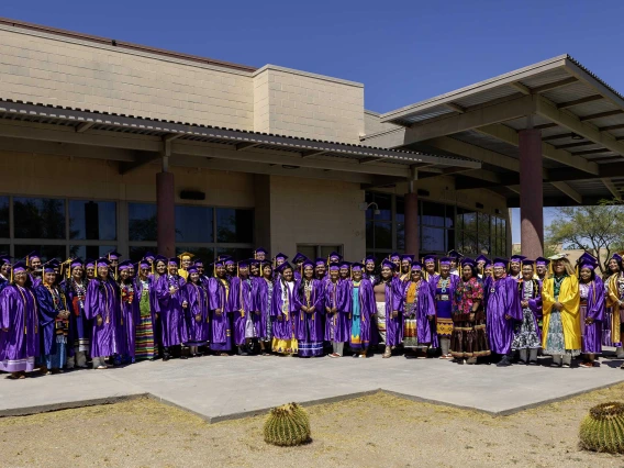 Graduates of the Tohono O’odham Community College Class of 2024 pose for a photo outside the college on a sunny day.