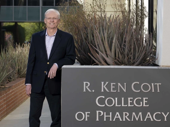 Rick G. Schnellmann, PhD, stands outside smiling as he leans on a sign that reads, “R. Ken Coint College of Pharmacy.”