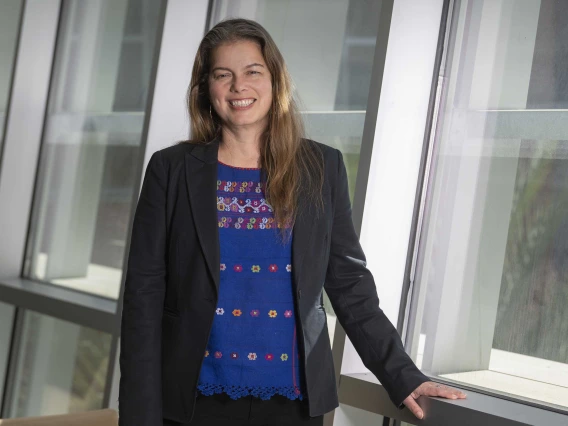 Portrait of Paloma Beamer, PhD, smiling as she stands next to a wall of windows. 