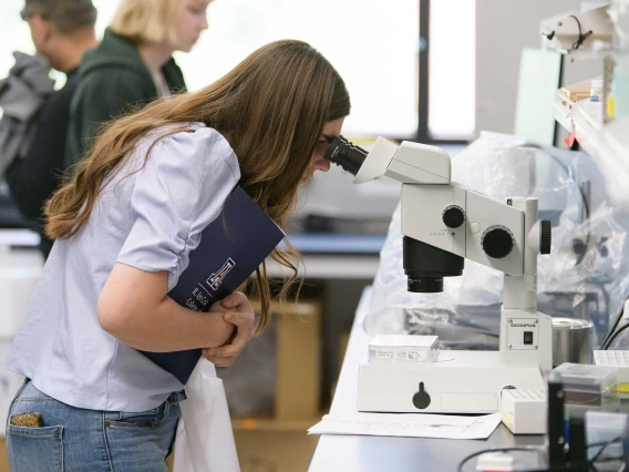 A student holds a folder to her chest while looking into a microscope.