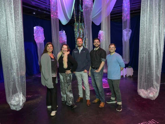 Five people post in the middle of the Hearing the Invisible immersive installation surrounded by columns of mesh fabric hanging from the ceiling