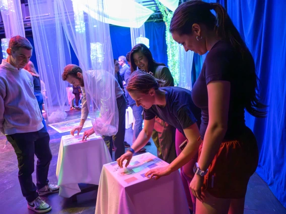 Three people standing around a small table watch as one person plays a card-marching memory game