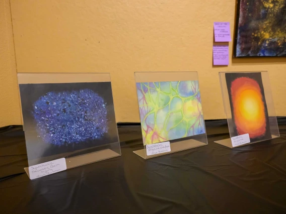 Three pieces of colorful artwork are displayed on a tabletop with a black tablecloth