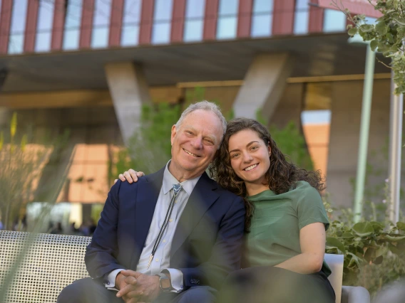 Geoffrey Rubin, MD, MBA, sits next to his daughter Elka Rubin on a bench outside. Their heads are leaned together, touching, and Elka’s arm is around her dad.